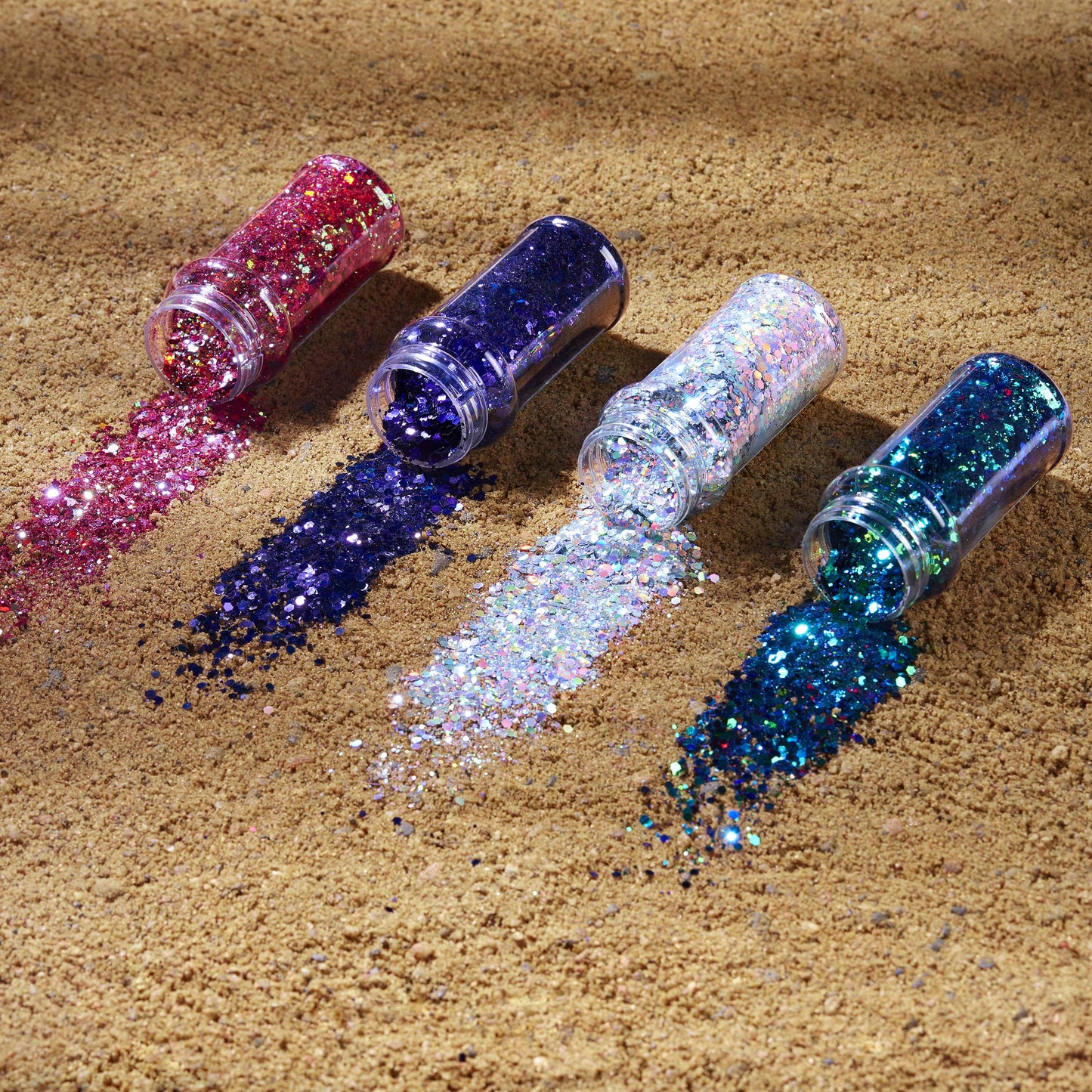Four bottles of glitter in pink, purple, silver, and blue on a sandy surface.