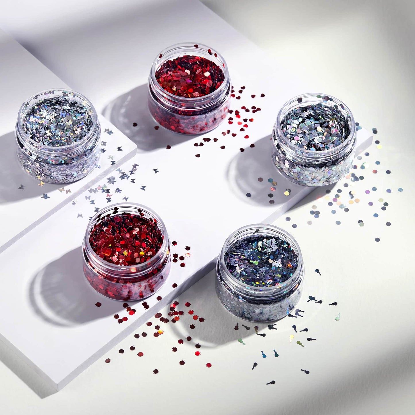 Five small jars of glitter on a white surface with scattered glitter.