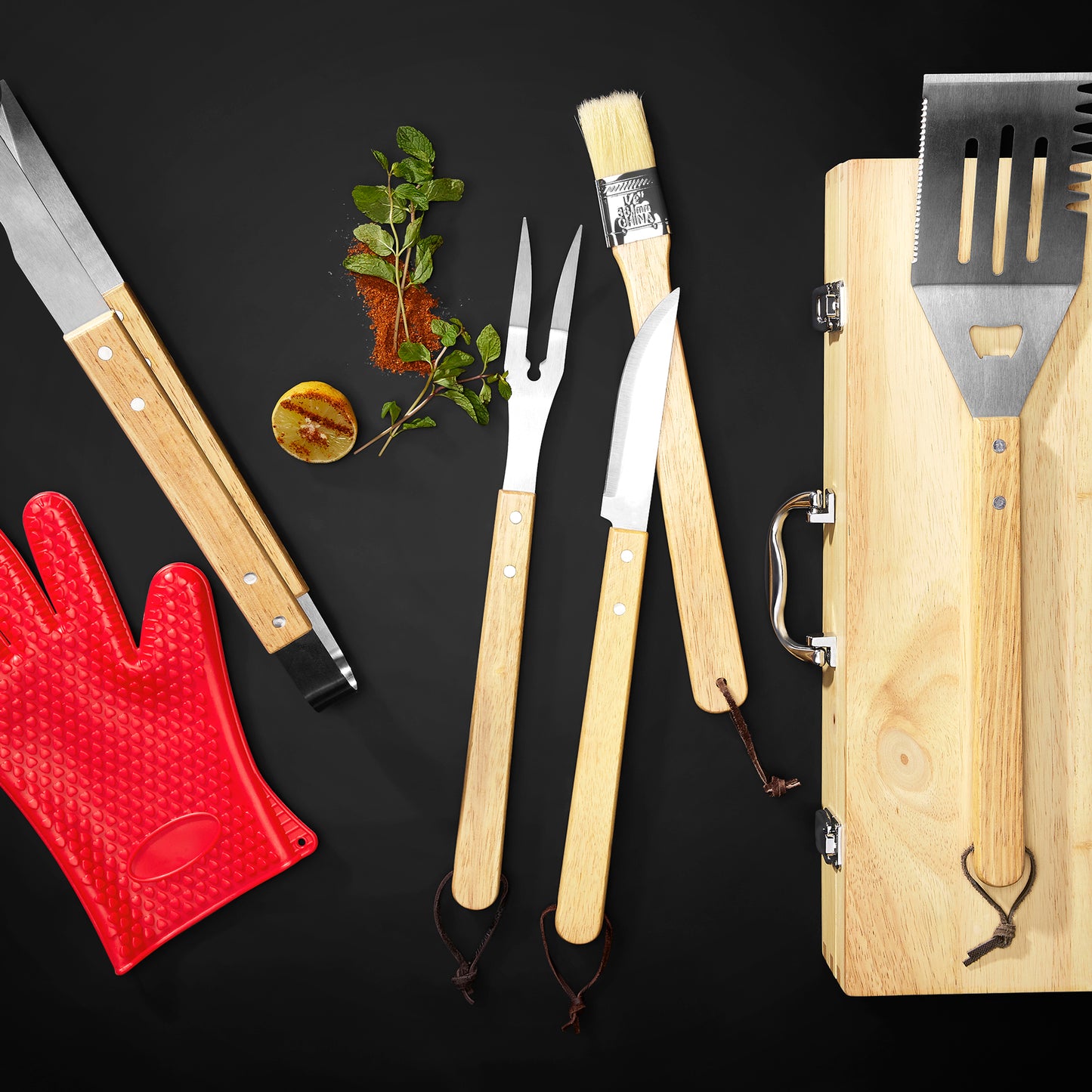 A barbecue tool set arranged on a black background, including a wooden-handled spatula, carving fork, brush, knife, and a wood carrying case. 