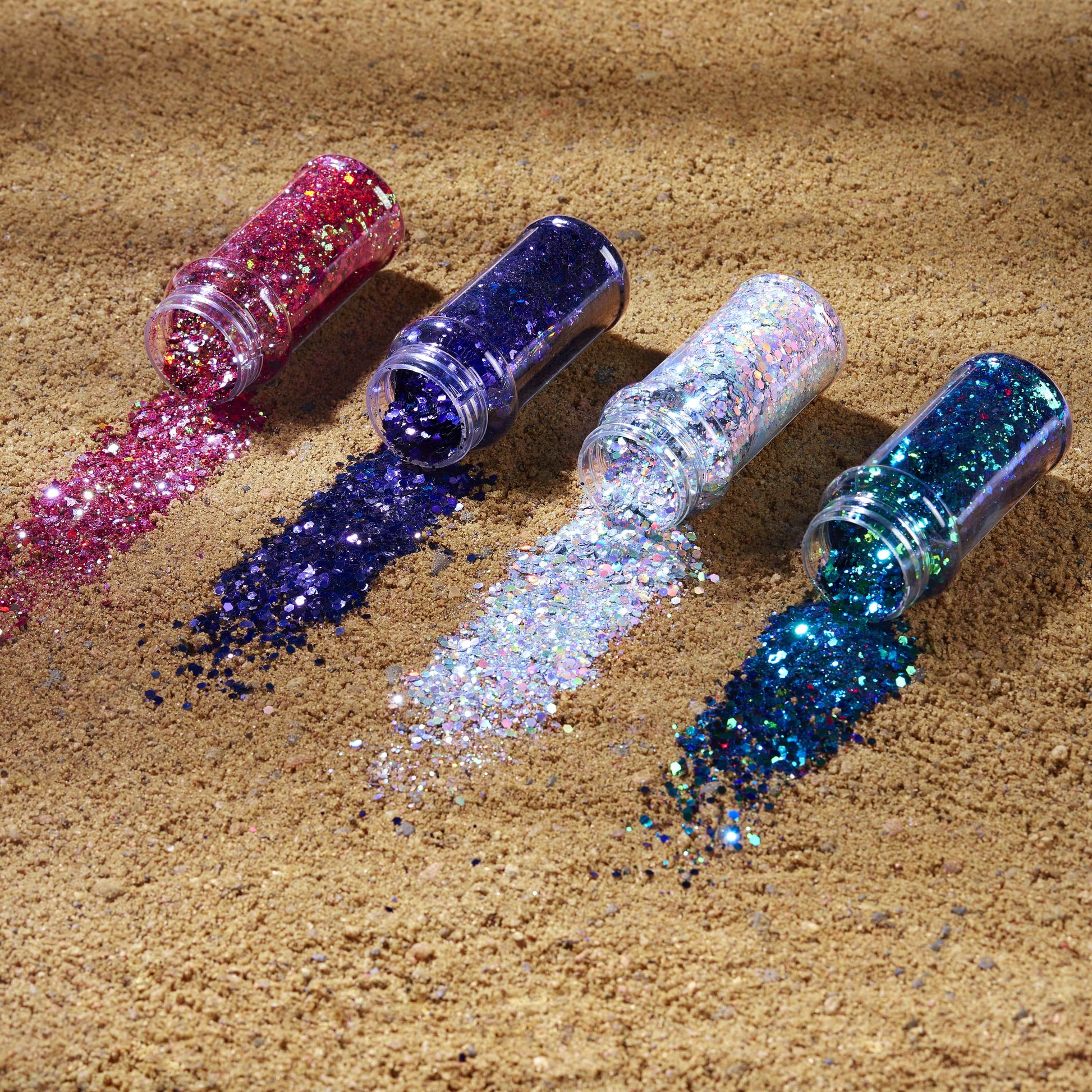 Four bottles of glitter in pink, purple, silver, and blue on a sandy surface.