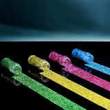 Four glittery sticks in green, gold, pink, and blue on a reflective surface with a dark background.
